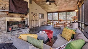 A cozy cabin interior with a stone fireplace, wooden ceiling, and a large window overlooking a forest.