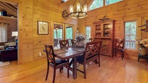 Interior de una cabaña de madera con una mesa de comedor, sillas, una araña y una zona de cocina.