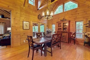 A wooden cabin interior with a dining table, chairs, a chandelier, and a kitchen area. - Glen Laurel Cabin (Cullowhee)