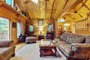 A log cabin interior with a wooden ceiling, a central fireplace, and a seating area with patterned sofas and a coffee table. - Glen Laurel Cabin (Cullowhee)