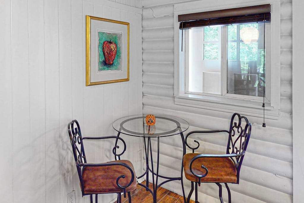 Una habitación con una mesa redonda de vidrio y dos sillas de metal, un cuadro enmarcado en la pared y una ventana con persianas.