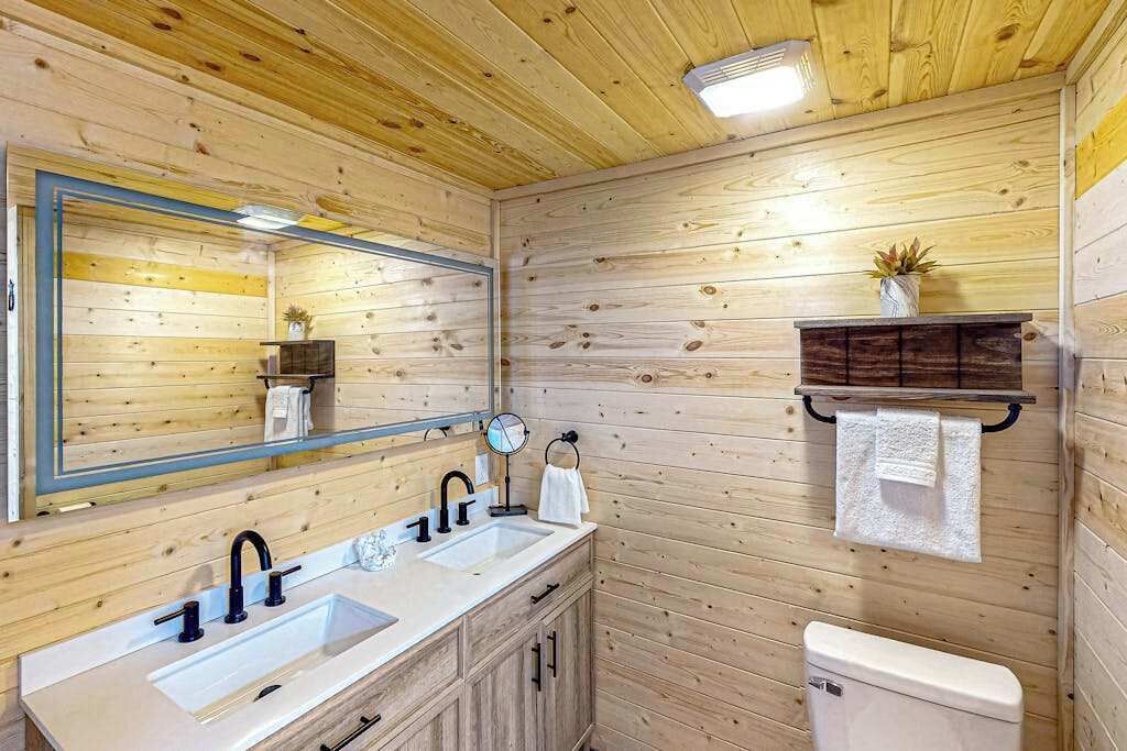 Een houten badkamer in een chalet, met een dubbele wastafel, toilet, spiegel en een plank met een plant.