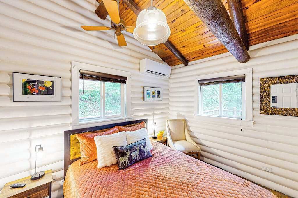 Een slaapkamer in een blokhut met een bed, twee ramen, een plafondventilator en een aan de muur bevestigde airconditioning.