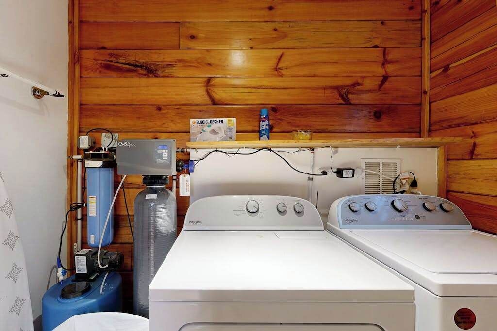 A laundry room with two Whirlpool washers, a water heater, and a shelf with a Black & Decker box.