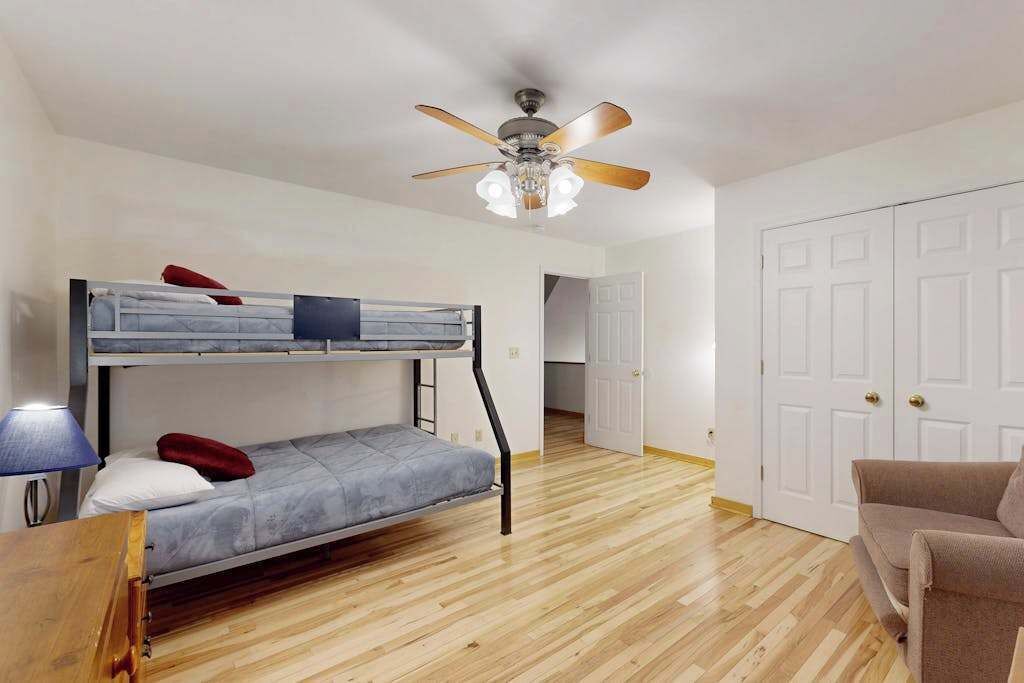 Une pièce avec un lit superposé, un ventilateur de plafond, une lampe, un plancher en bois et une porte.