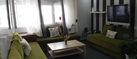 A living room with a green sofa, a wooden coffee table, a TV mounted on the wall, and a window with curtains.