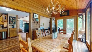 Un interior de madera con una mesa de comedor, sillas, una araña y una vista por la puerta.