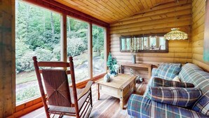 Interior de una cabaña de madera con un ventanal grande, una mesa de madera, un sofá y una lámpara colgante.