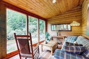 A wooden cabin interior with a large window, a wooden table, a sofa, and a hanging light fixture. - Misty Falls Hideaway (Cullowhee)