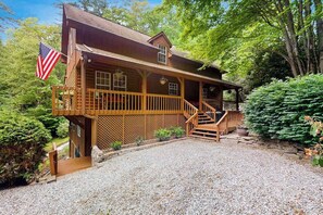 A wooden cabin with a porch, a flag, and a gravel driveway. - Misty Falls Hideaway (Cullowhee)