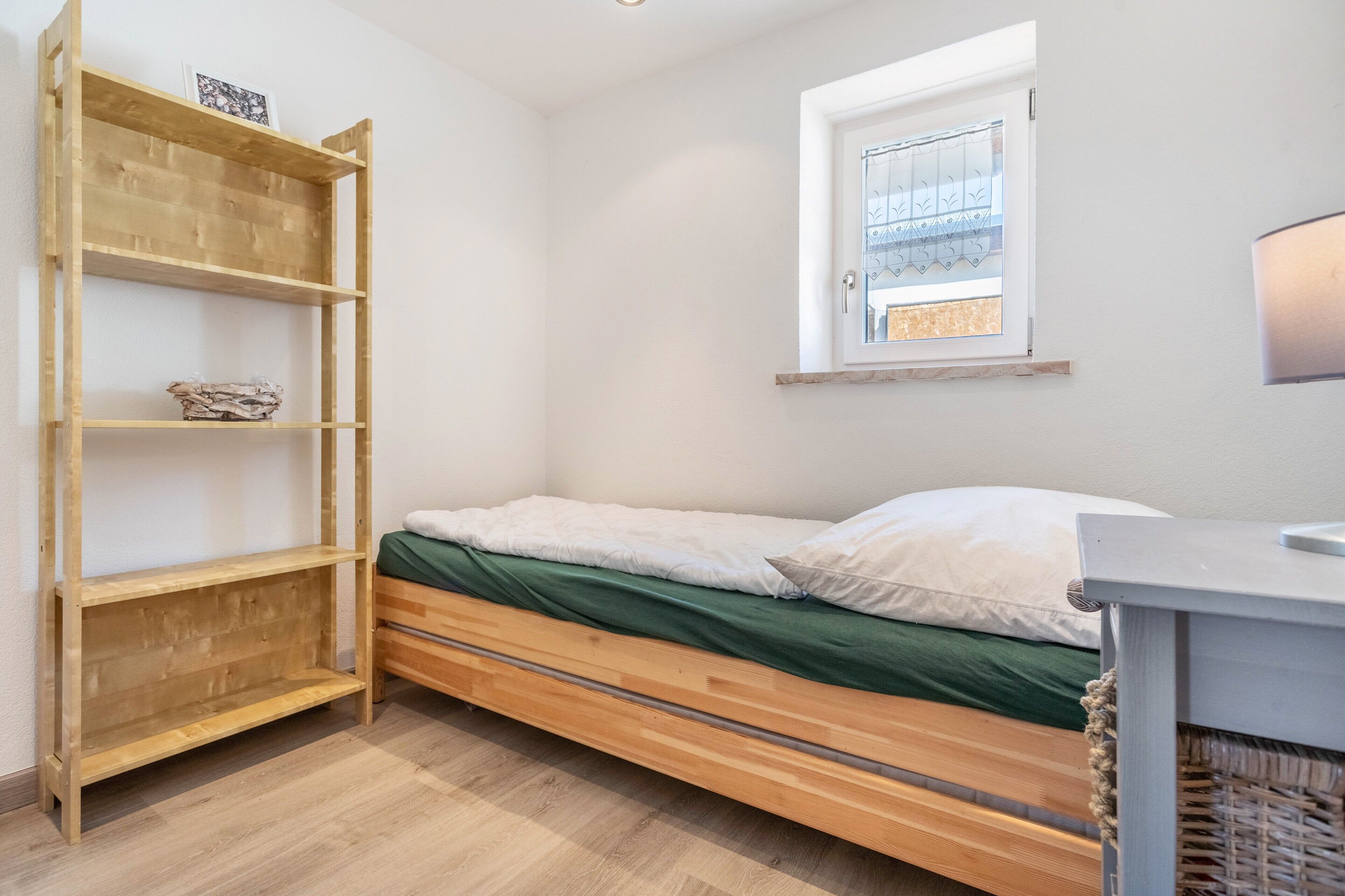 A single bed with a green bedspread, a wooden bookshelf with a picture on the top shelf, and a window with a metal grill.