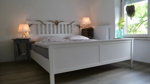 A white bed with a headboard, two bedside lamps, a nightstand, and a window with a view of greenery. - Holiday Home for Up to 6 Guests (70 m²) in Trent (Rügen) (Trent)