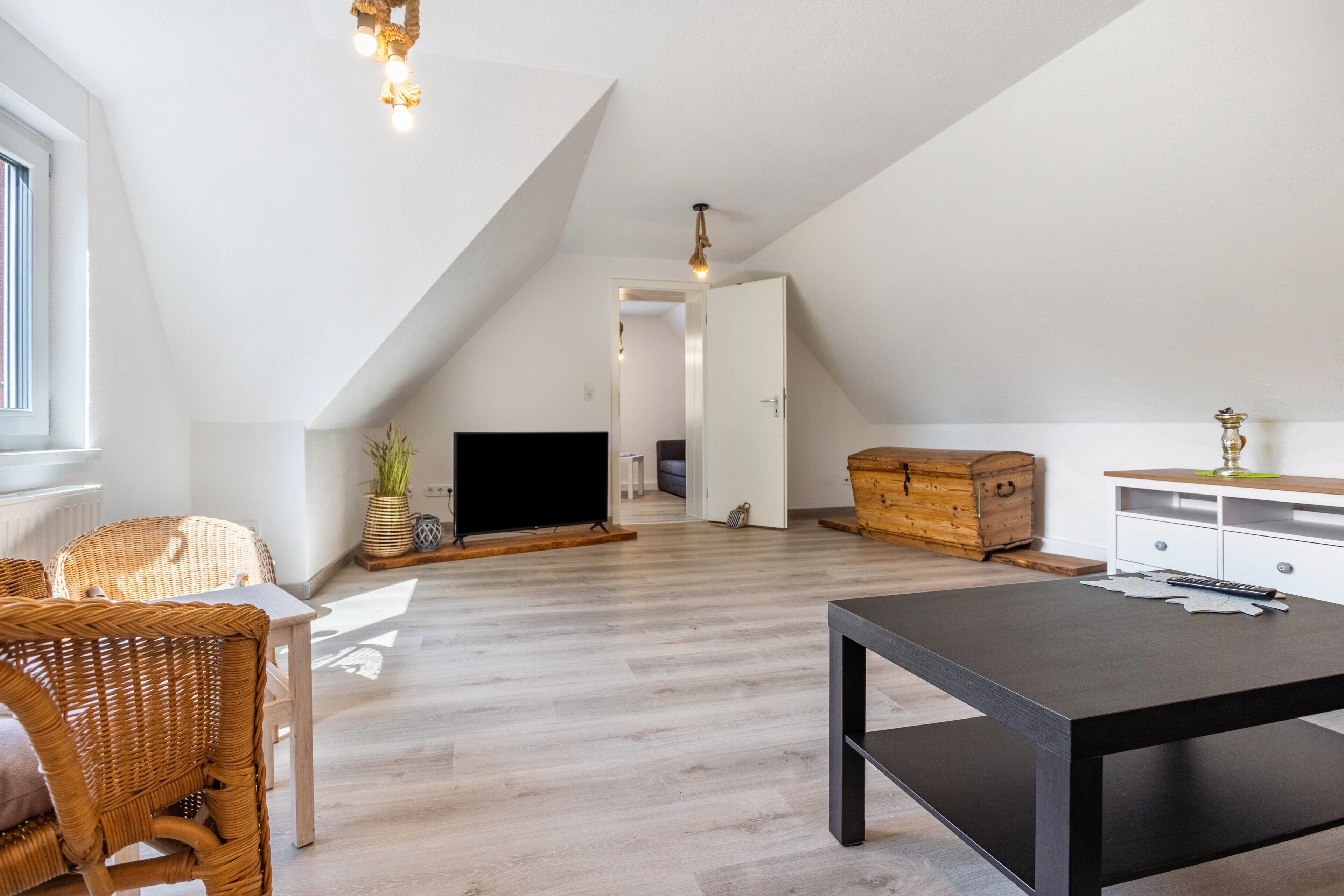 A spacious loft bedroom with a sloped ceiling, a large window, a flat-screen TV, a wooden chest, and a dining table.