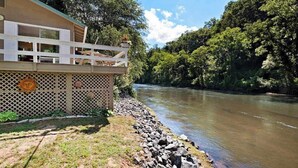 A riverside building with a balcony, a wooden fence, and a rocky riverbank.