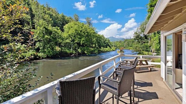 A deck with wicker chairs overlooking a river, surrounded by lush greenery and a clear sky.
