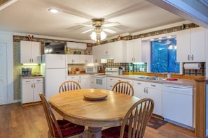 Une cuisine compacte avec des meubles blancs, une table à manger avec des chaises, un ventilateur de plafond et une fenêtre donnant sur un paysage verdoyant.