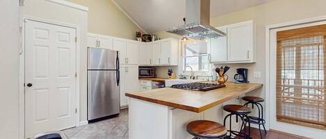 A kitchen with white cabinets, a stainless steel refrigerator, a wooden island with stools, and a window with blinds.