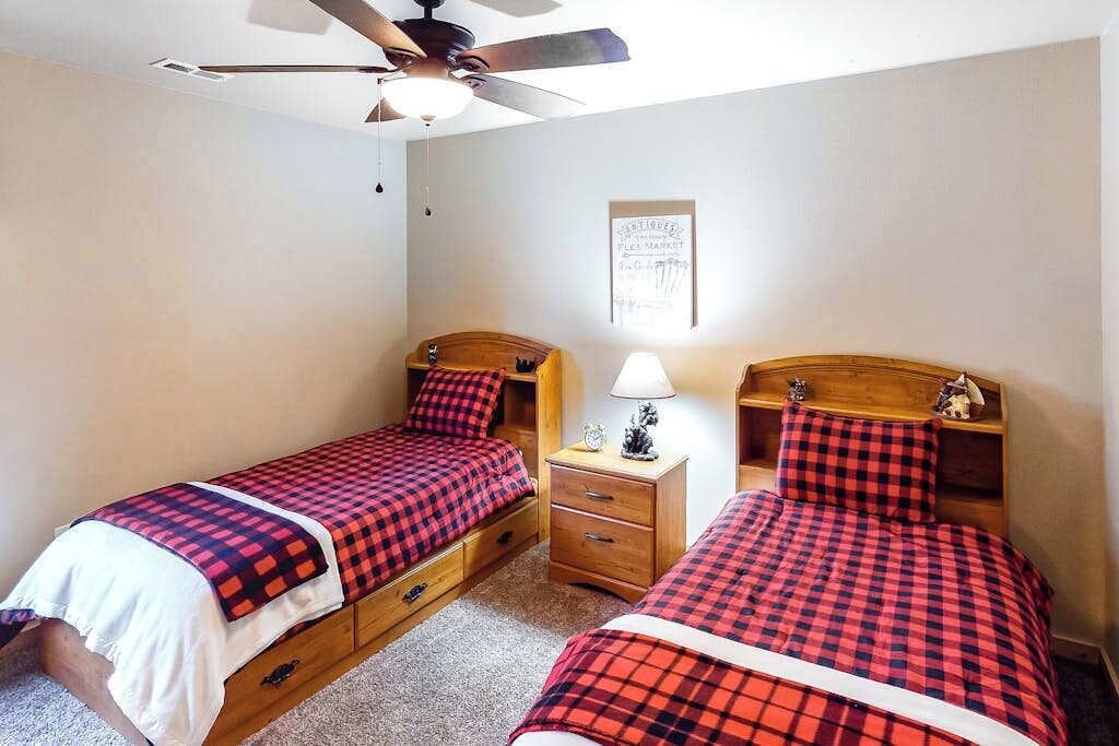 Une chambre avec deux lits simples, des têtes de lit en bois, un ventilateur de plafond, une table de chevet avec une lampe et un tableau encadré au mur.