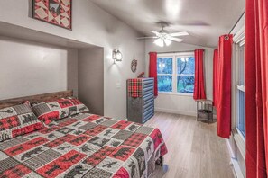 Un dormitorio con una cama, un ventilador de techo, una ventana con cortinas rojas y una mesita con una lámpara.