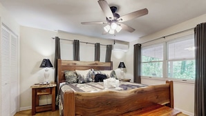 A bedroom with a wooden bed, a ceiling fan, a bedside table, a bench, and a window with curtains.