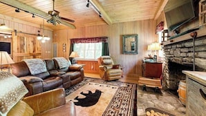 A cozy cabin interior with a leather sofa, a stone fireplace, and a television mounted on the wall.
