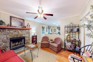 A cozy living room with a stone fireplace, a ceiling fan, and a bookshelf. - The Cottage on Chowan (Highlands)