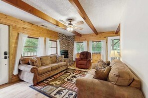 A log cabin interior with a fireplace, wooden beams, a ceiling fan, and a large rug with animal prints. - Hollow's End (Whittier)