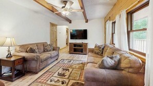 Ein Wohnzimmer mit zwei Sofas, einem Fernseher, Holzbalken, einem Deckenventilator und einem Fenster mit Vorhängen.