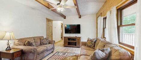 A living room with two sofas, a TV, wooden beams, a ceiling fan, and a window with curtains.