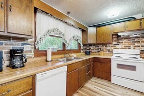 A compact kitchen with wooden cabinets, a white dishwasher, a stove, and a coffee maker. - Hollow's End (Whittier)