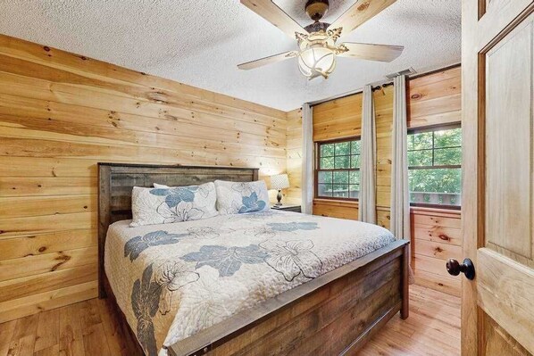 A wooden-paneled bedroom with a bed, a ceiling fan, and a window. - Hollow's End (Whittier)