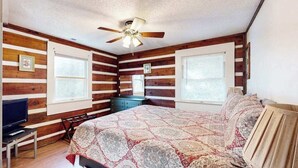 A bedroom with wooden walls, a ceiling fan, a bed with a patterned bedspread, a television on a stand, and a window with blinds. - Family Jewel (Whittier)