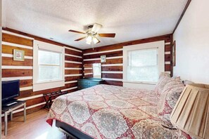 Ein Schlafzimmer mit Holzwänden, einem Deckenventilator, einem Bett mit gemustertem Bettbezug, einem Fernseher auf einem Standfuß und einem Fenster mit Jalousien.