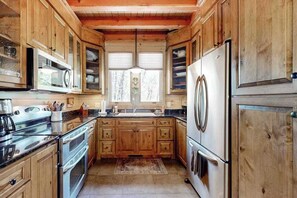 A kitchen with wooden cabinets, stainless steel appliances, and a window with a view of trees. - Chic Highland Cabin (Highlands)