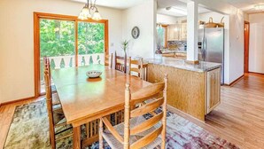 Un comedor con una mesa y sillas de madera, una cocina con refrigerador, y vista a la vegetación por una ventana.
