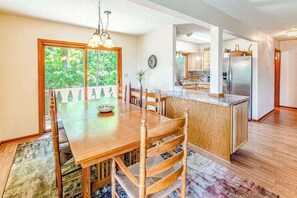 A dining area with a wooden table and chairs, a kitchen with a refrigerator, and a view of greenery through a window. - Beautiful home with hot tub, firepit, deck, & central AC (Whittier)