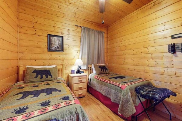 Een blokhut met twee houten bedden, een nachtkastje, een lamp, een stoel en een raam met gordijnen.