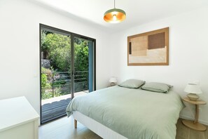 Un dormitorio con cama, mesita de noche, lámpara, un cuadro y vistas a la vegetación a través de una puerta corredera.