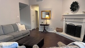 A living room with a grey sofa, a patterned armchair, a dark ottoman, a fireplace, and a mirror on the wall. - Appomattox Family House (College Station)