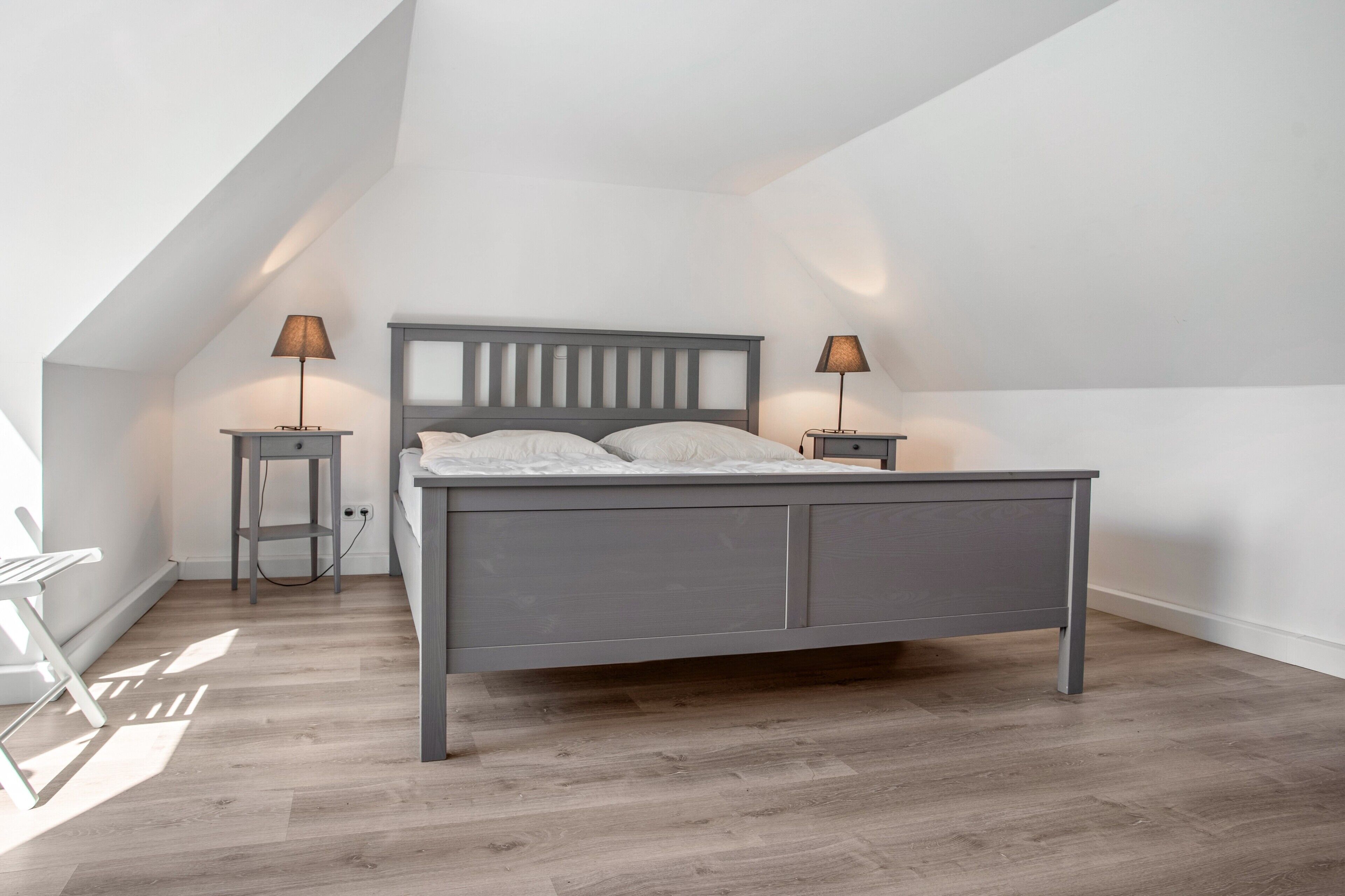 A bedroom with a sloped ceiling, a bed with a grey headboard, two bedside tables with lamps, and a wooden floor.