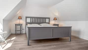 Une chambre avec un plafond en pente, un lit doté d’une tête de lit grise, deux tables de chevet avec des lampes et un plancher en bois.