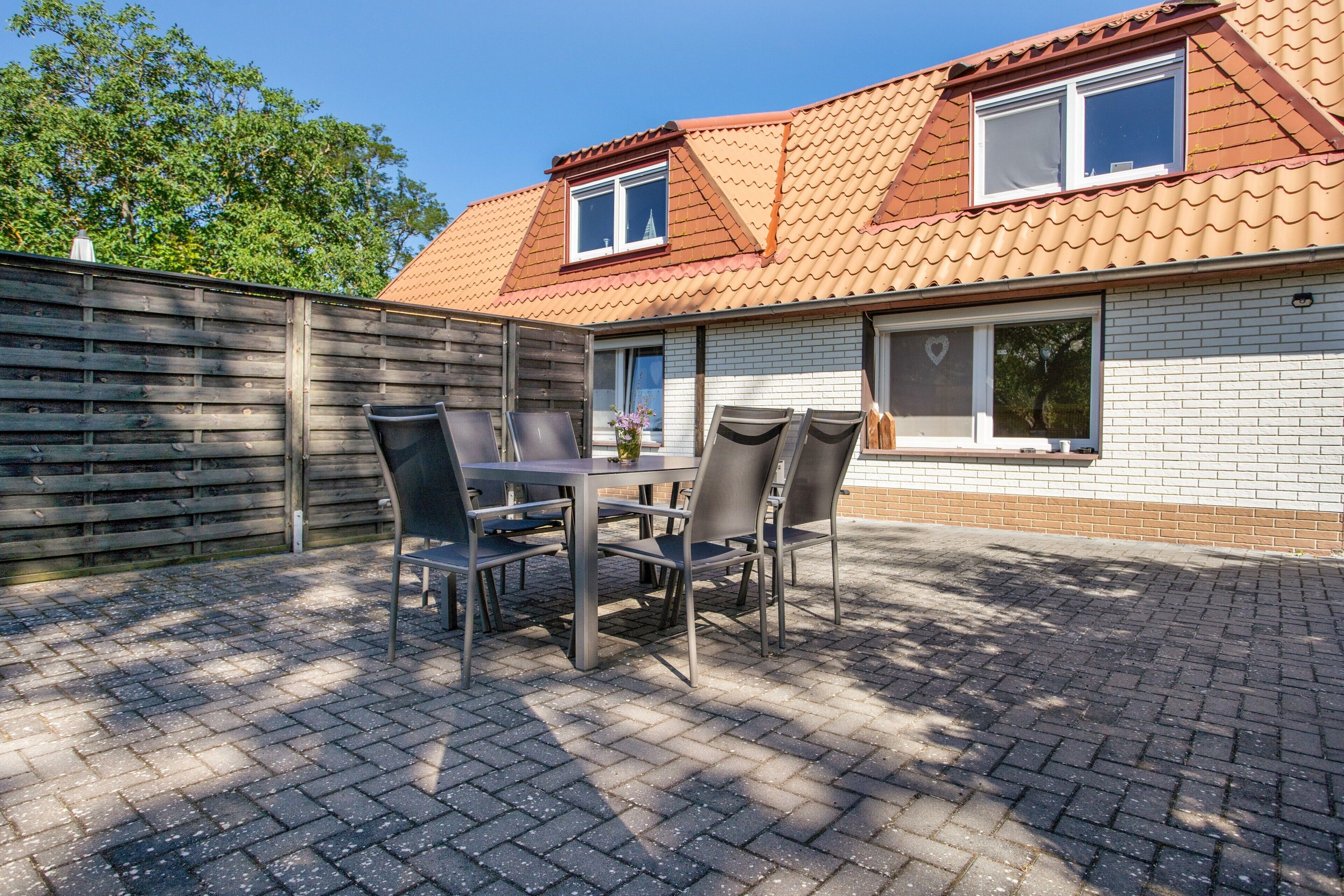 Une terrasse avec une table et des chaises, une clôture en bois et une maison avec un toit en tuiles et des fenêtres.