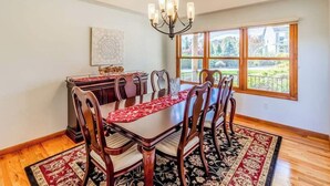 A dining room with a wooden table, six chairs, a chandelier, a red and black patterned rug, and a window with a view of greenery. - Everview (Waynesville)