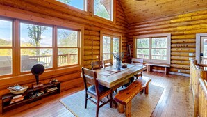 Ein Holzblockhaus-Interieur mit einem Holzesstisch, Stühlen, einer Bank und großen Fenstern, die einen Blick ins Freie bieten.
