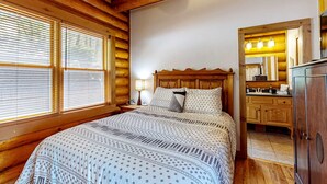 Ein Schlafzimmer in einer Blockhütte mit Bett, Holz-Kopfschild, Nachttischlampe und Blick ins Badezimmer.