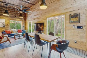 Un intérieur moderne avec un plafond en bois, un coin repas comprenant une table et des chaises en bois, un canapé agrémenté de coussins colorés et un téléviseur fixé au mur.