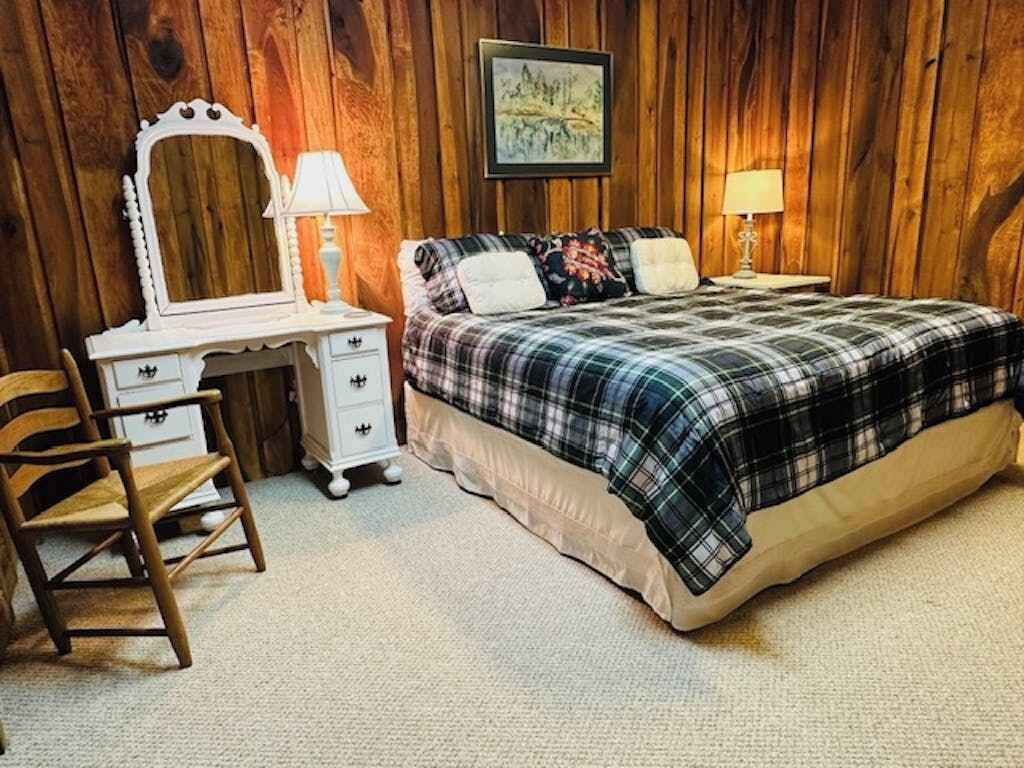 Een slaapkamer met een houten wand, een bed met een geruite plaid, een dressoir met spiegel en twee bedlampjes.