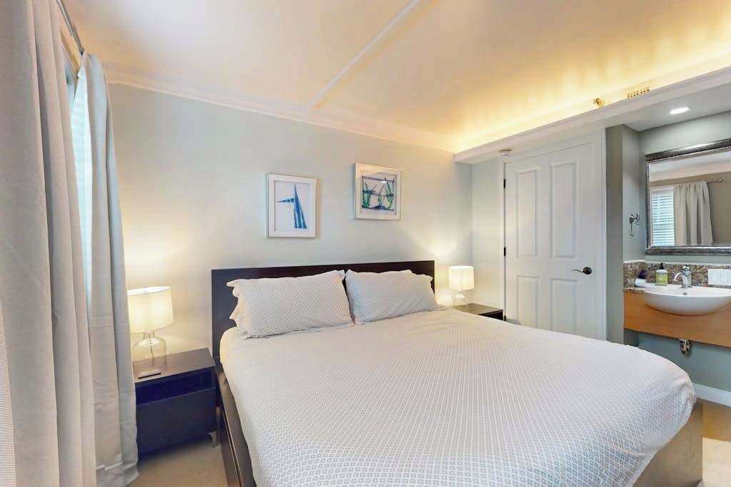 A hotel room with a bed, bedside lamps, a nightstand, a bathroom with a sink and mirror, and two framed pictures on the wall.