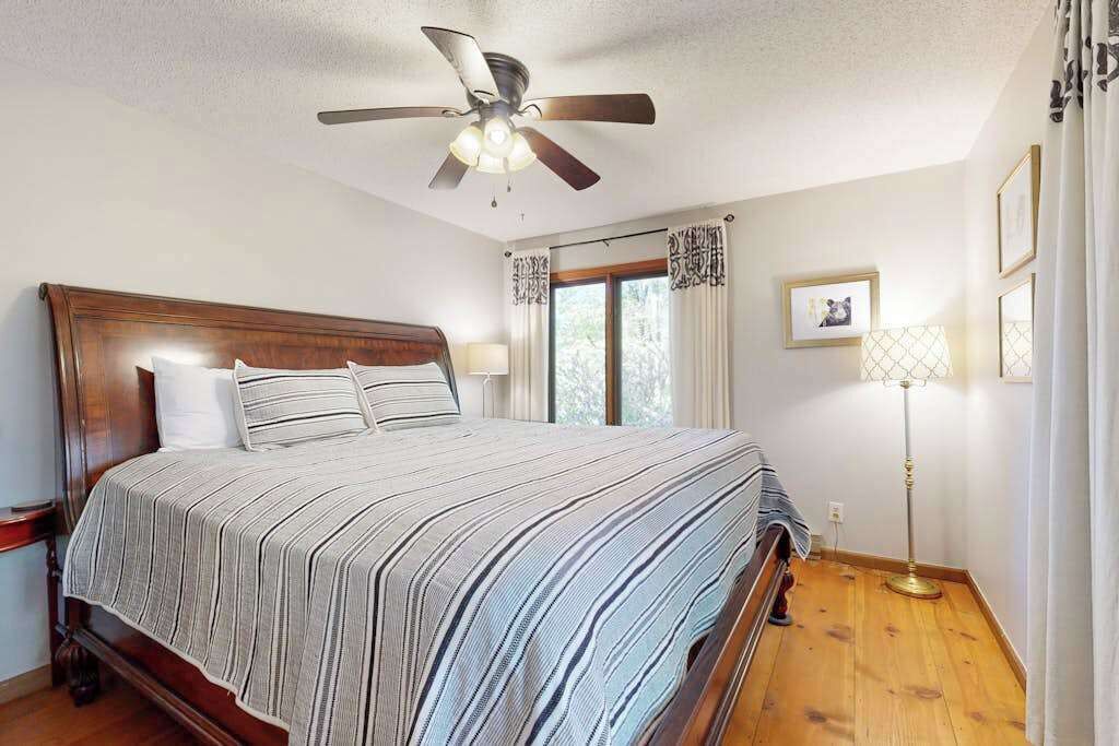 Un dormitorio con cama de madera, ventilador de techo, lámpara de pie y ventana con cortinas.