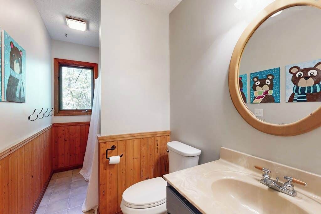 Une salle de bain avec un meuble-lavabo en bois, des toilettes blanches, une fenêtre et des œuvres d’art sur le thème des ours.